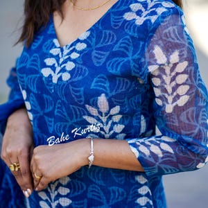 May include: A blue and white cotton kurta with a floral print. The kurta has a V-neck and three-quarter sleeves. The fabric is sheer and has a slightly textured look. The text "Bahe Kurtis" is visible on the sleeve.