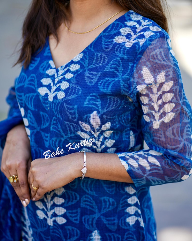 May include: A blue and white cotton kurta with a floral print. The kurta has a V-neck and three-quarter sleeves. The fabric is sheer and has a slightly textured look. The text "Bahe Kurtis" is visible on the sleeve.