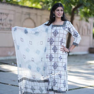 May include: A white cotton kurta with a geometric print and a matching white palazzo pant. The kurta has a square neckline and three-quarter sleeves. The outfit is paired with a white sheer dupatta with a geometric print. The model is wearing the outfit and standing on a sidewalk.