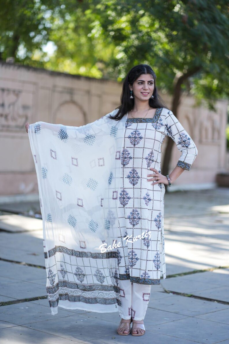 May include: A white cotton kurta with a geometric print and a matching white palazzo pant. The kurta has a square neckline and three-quarter sleeves. The outfit is paired with a white sheer dupatta with a geometric print. The model is wearing the outfit and standing on a sidewalk.
