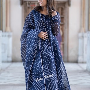 May include: A woman wearing a blue and white patterned dress with a matching dupatta. The dress has a long, flowing skirt and a fitted bodice. The dupatta is draped over her shoulders and is secured at her waist. The woman is standing in front of a building with a tiled floor.