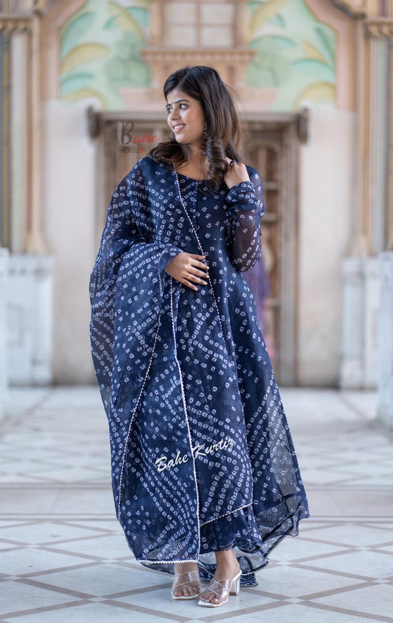 May include: A woman wearing a blue and white patterned dress with a matching dupatta. The dress has a long, flowing skirt and a fitted bodice. The dupatta is draped over her shoulders and is secured at her waist. The woman is standing in front of a building with a tiled floor.