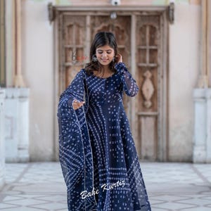 May include: A woman wearing a blue and white patterned dress with a matching dupatta. The dress has a long, flowing skirt and a fitted bodice. The woman is standing in front of a building with a wooden door.