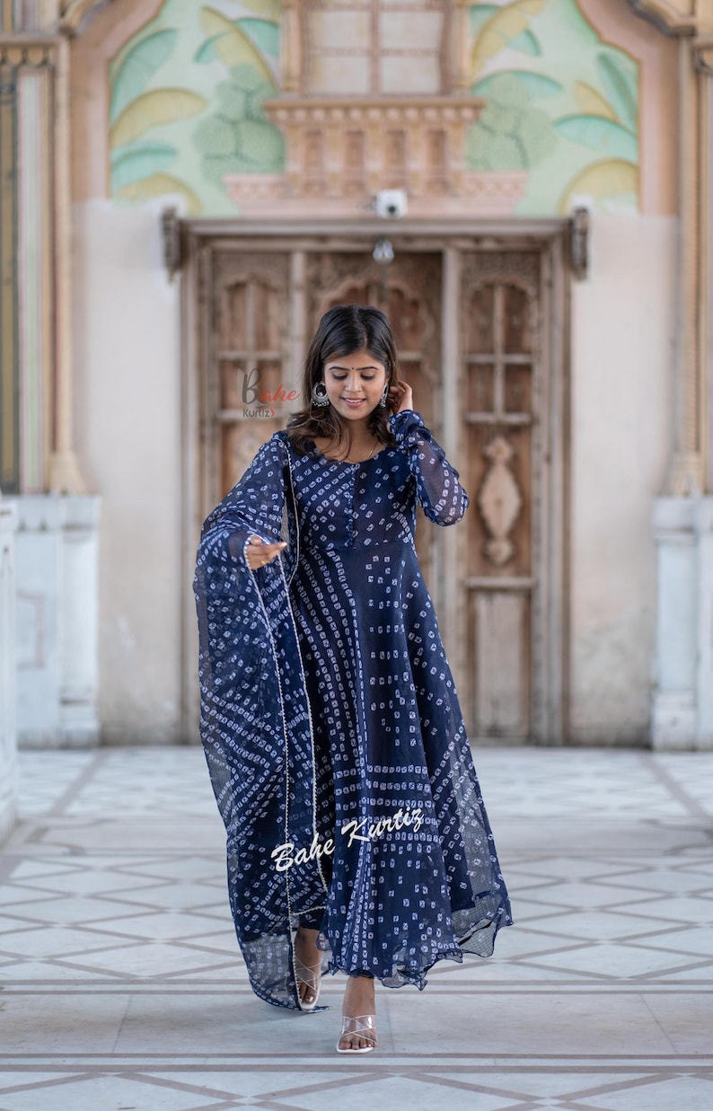 May include: A woman wearing a blue and white patterned dress with a matching dupatta. The dress has a long, flowing skirt and a fitted bodice. The woman is standing in front of a building with a wooden door.