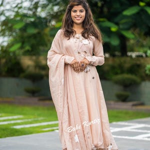 May include: A light peach-colored long-sleeved dress featuring a subtle floral pattern in white and brown tones. The garment includes a matching draped shawl or dupatta and displays the text Bahe Kurtiz in white script on the lower front section.