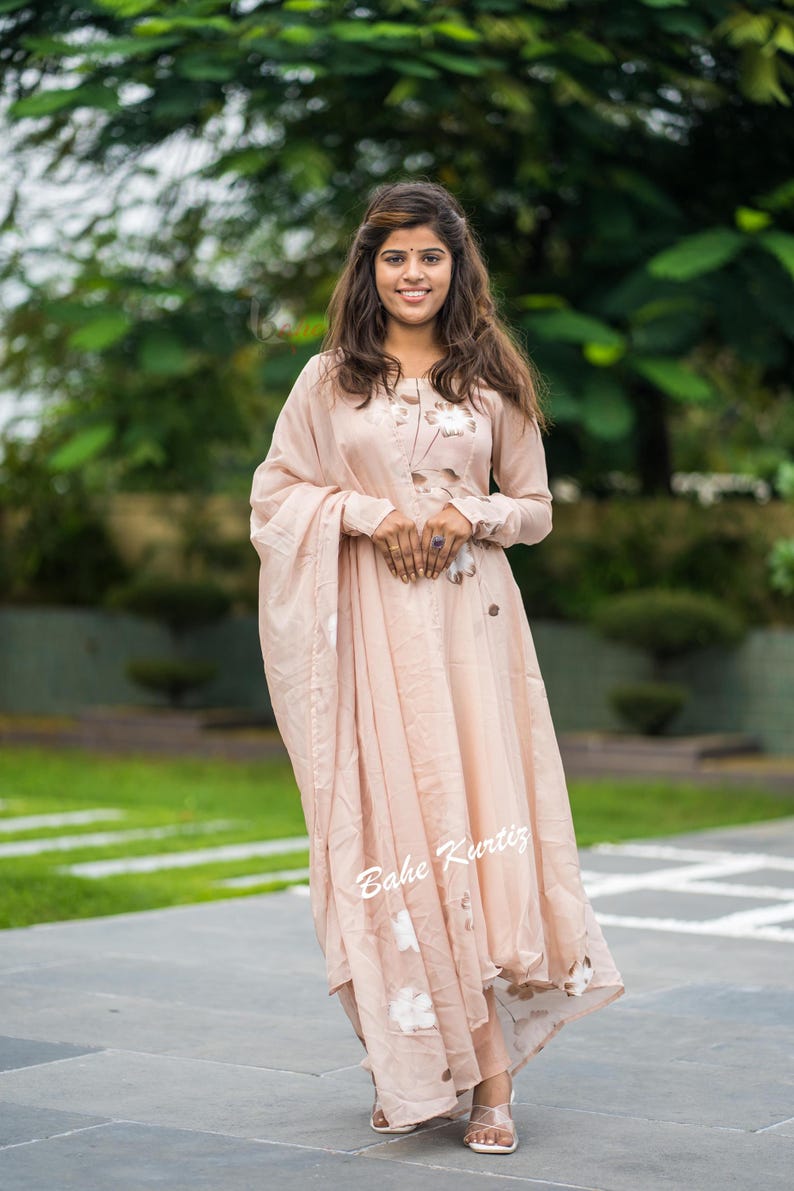 May include: A light peach-colored long-sleeved dress featuring a subtle floral pattern in white and brown tones. The garment includes a matching draped shawl or dupatta and displays the text Bahe Kurtiz in white script on the lower front section.