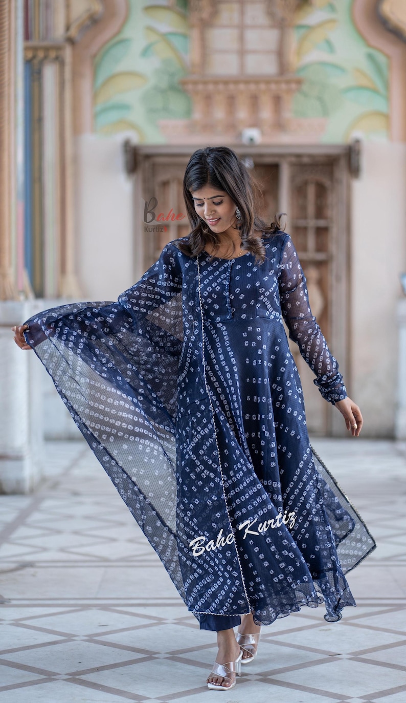May include: A navy blue long-sleeved dress with a white print. The dress has a flowing silhouette and is paired with a matching dupatta. The dress is adorned with pearl embellishments. The model is wearing silver sandals.