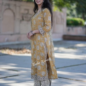 May include: A woman wearing a yellow and white printed kurta with a matching palazzo pant. The kurta has a black border and the palazzo pant has a black and white geometric print. The woman is standing in front of a green bush.