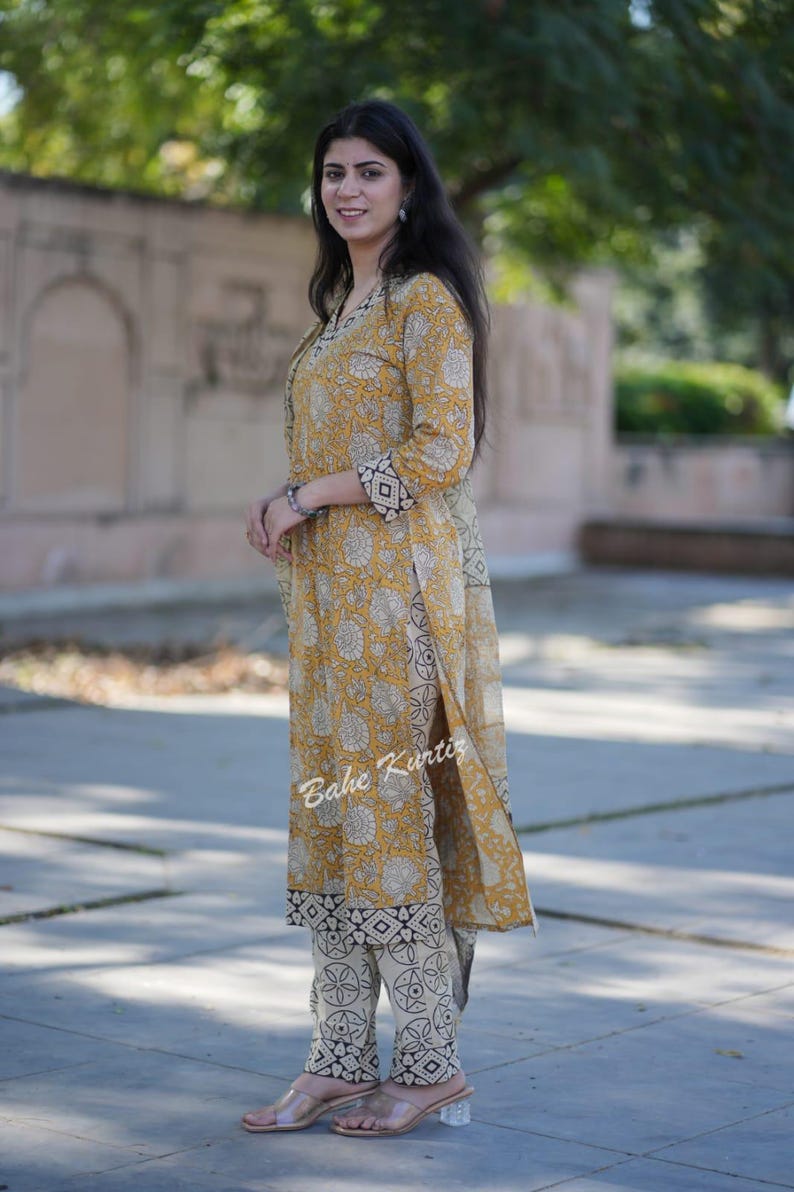 May include: A woman wearing a yellow and white printed kurta with a matching palazzo pant. The kurta has a black border and the palazzo pant has a black and white geometric print. The woman is standing in front of a green bush.