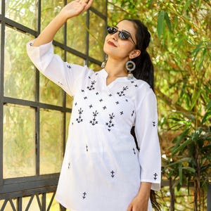 May include: A white cotton top with black embroidered anchors and crosses. The top has a V-neck and three-quarter sleeves. The model is wearing blue jeans and sunglasses.