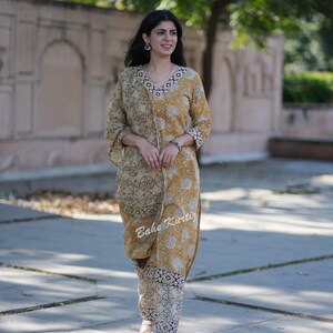 May include: A woman wearing a yellow and white printed kurta with a matching printed palazzo pants. The kurta has a V-neckline and a black and white geometric pattern on the neckline. The palazzo pants have a black and white geometric pattern. The woman is walking on a paved path.