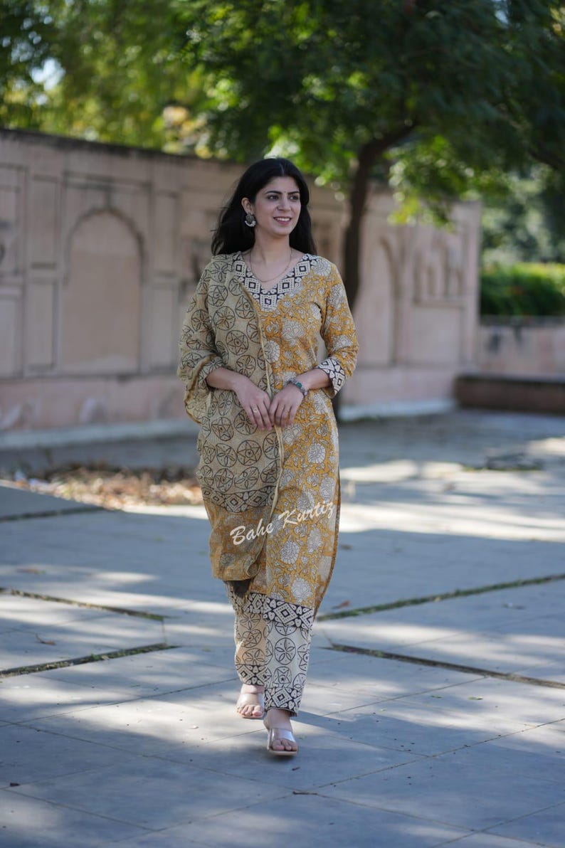 May include: A woman wearing a yellow and white printed kurta with a matching printed palazzo pants. The kurta has a V-neckline and a black and white geometric pattern on the neckline. The palazzo pants have a black and white geometric pattern. The woman is walking on a paved path.