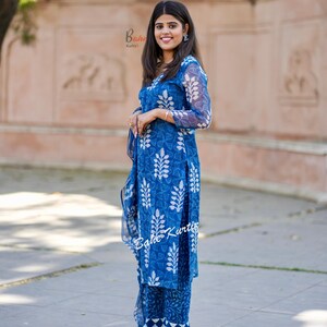 May include: A woman wearing a blue and white printed kurta and matching pants. The kurta has a V-neckline and long sleeves. The pants are straight-legged and have a white trim at the bottom. The woman is standing in front of a brick wall.