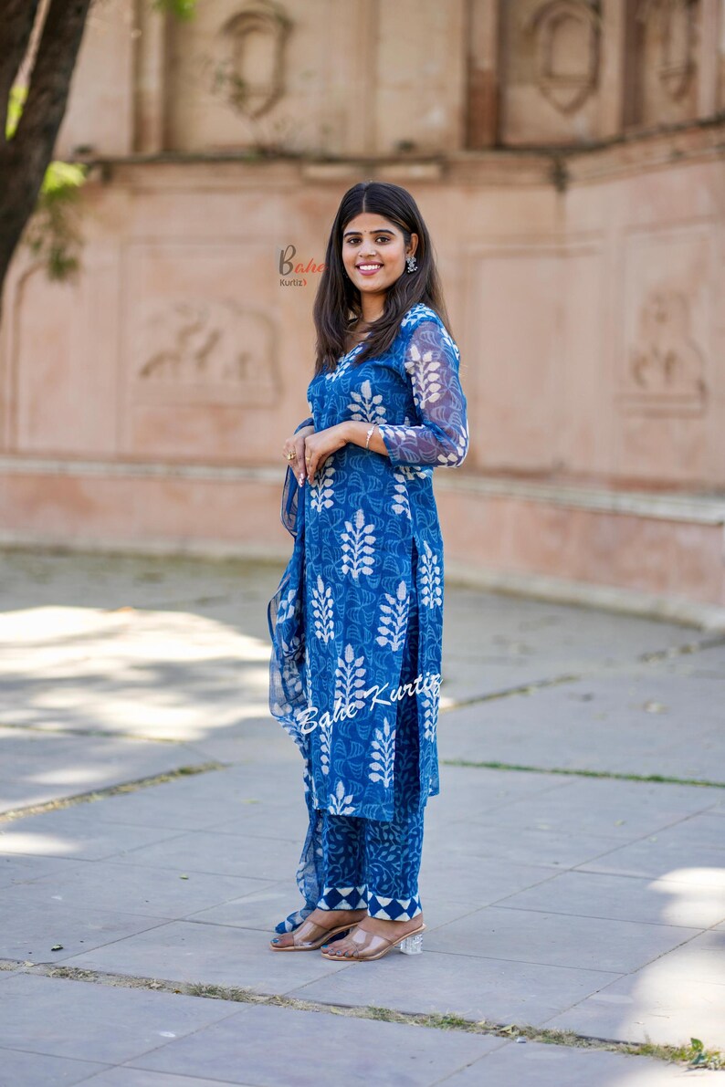 May include: A woman wearing a blue and white printed kurta and matching pants. The kurta has a V-neckline and long sleeves. The pants are straight-legged and have a white trim at the bottom. The woman is standing in front of a brick wall.