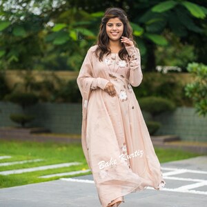 May include: A light peach-colored long-sleeved dress featuring a subtle floral pattern and a matching sheer dupatta draped over the shoulder. The garment has a round neckline and a flowing silhouette. The text Bahe Kurtiz is visible on the fabric.