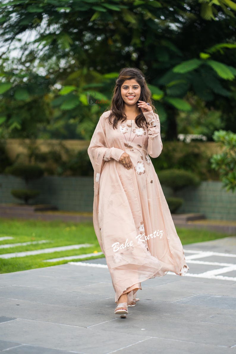 May include: A light peach-colored long-sleeved dress featuring a subtle floral pattern and a matching sheer dupatta draped over the shoulder. The garment has a round neckline and a flowing silhouette. The text Bahe Kurtiz is visible on the fabric.