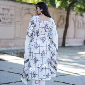 May include: A white and blue patterned salwar kameez with a square neckline and a dupatta. The salwar kameez has a checkered pattern with floral designs. The dupatta is a sheer white fabric with a floral border.