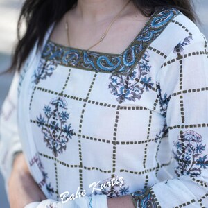 May include: White cotton kurta with a green and blue floral print. The kurta has a square neckline with a green and blue embroidered trim. The sleeves are three-quarter length and also have a green and blue embroidered trim. The kurta is paired with a white dupatta. The text "Bahe Kurtiz" is visible on the kurta.