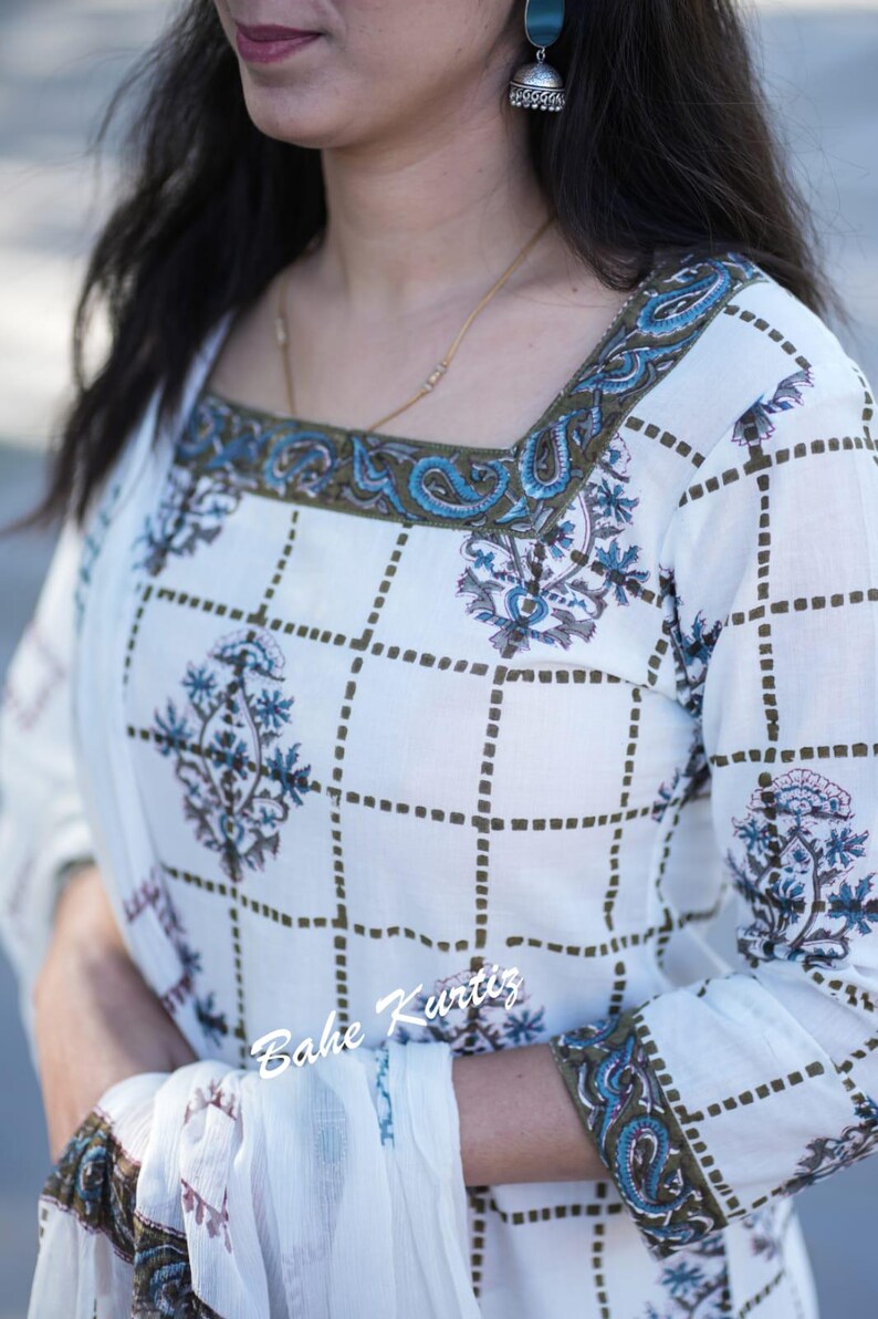 May include: White cotton kurta with a green and blue floral print. The kurta has a square neckline with a green and blue embroidered trim. The sleeves are three-quarter length and also have a green and blue embroidered trim. The kurta is paired with a white dupatta. The text "Bahe Kurtiz" is visible on the kurta.