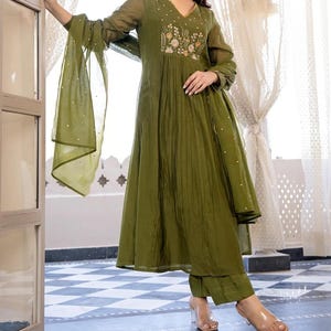 May include: Olive green embroidered tunic and matching trousers with a sheer scarf. The tunic features a V-neck and floral embroidery. The outfit is made of a lightweight fabric, suitable for warm weather.