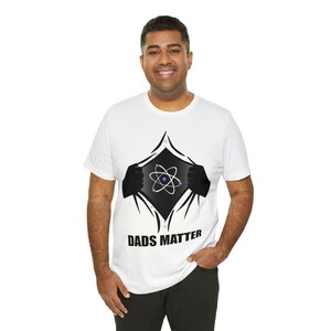 May include: White t-shirt featuring a graphic design of a superhero-style reveal, with the text "DADS MATTER" below a stylized atom symbol. The design is printed on a white t-shirt.