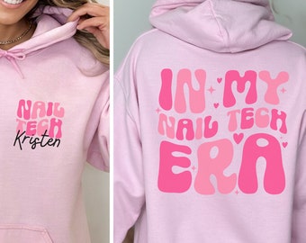 May include: Light pink hoodie with the text "NAIL TECH Kristen" on the front and "IN MY NAIL TECH ERA" on the back. The text is in shades of pink. The hoodie has a hood and a front pocket.