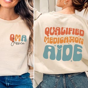 May include: Beige sweatshirt with the text "OMA Jessica" in orange and teal on the front. The back of the sweatshirt features the words "QUALIFIED MEDICATION AIDE" in orange and teal, with a retro font.