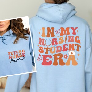 May include: Light blue hoodie with the text "In My Nursing Student Era" in colourful retro font. The text is decorated with a butterfly, a stethoscope, a syringe, a flower, and a nurse's cap. The hoodie has a drawstring hood and a kangaroo pocket.