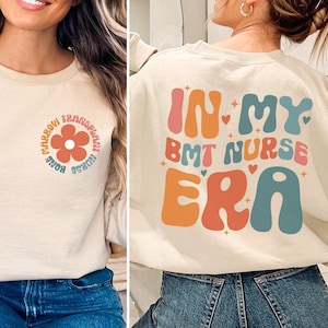 May include: A beige sweatshirt with a graphic design on the front and back. The front design features a flower with the text "Bone Marrow Transplant Nurse" surrounding it. The back design features the text "In My BMT Nurse Era" in a retro font with hearts and stars.