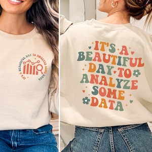 以下が含まれることがあります： 薄いベージュのトレーナーで、前面と背面にグラフィックデザインが施されています。前面のデザインは、棒グラフと虫眼鏡が描かれた円形です。背面のデザインは、「データ分析をするのに最適な日」というカラフルでスタイリッシュなテキストです。