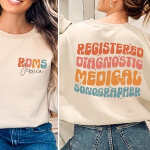 Könnte beinhalten: Ein beiger Pullover mit einer bunten Grafik auf der Vorderseite, die "RDMS Jessica" sagt, und einer bunten Grafik auf der Rückseite, die "Registered Diagnostic Medical Sonographer" sagt.