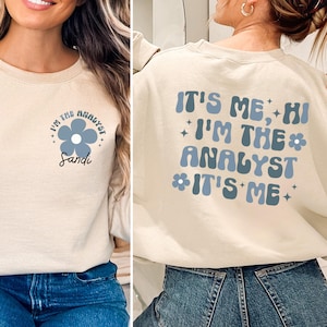 Może przedstawiać: Kremowa bluza z napisem "I'M THE ANALYST" i niebieskim kwiatem z przodu. Tył bluzy zawiera napis "IT'S ME, HI I'M THE ANALYST IT'S ME" w kolorze niebieskim. Bluza ma swobodny styl.
