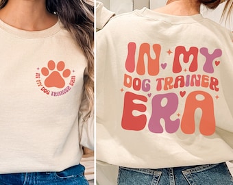 May include: A beige sweatshirt with the text "In My Dog Trainer Era" in a retro font with pink, orange, and purple colors. The text is on the back of the sweatshirt. The front of the sweatshirt has a paw print with the same text in a smaller font.
