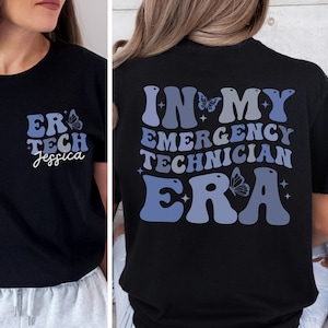 Könnte beinhalten: Schwarzes T-Shirt mit einer blauen und weißen Grafik, auf der "In My Emergency Technician Era" mit einem Schmetterlingsdesign steht. Die Vorderseite des Shirts hat eine kleinere Grafik, auf der "ER Tech Jessica" mit einem Schmetterlingsdesign steht.