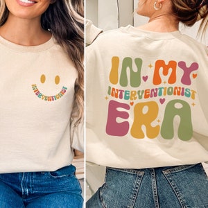May include: A beige sweatshirt with a smiley face on the front and the text "In My Interventionist Era" on the back. The text is in a retro font with colorful letters and stars.