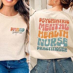 Op de afbeelding: Een beige sweatshirt met een retro-grafisch ontwerp met de tekst "Psychiatric Mental Health Nurse Practitioner". De grafiek is in tinten blauw, oranje en roze. Het sweatshirt wordt gedragen door een persoon met lang bruin haar.