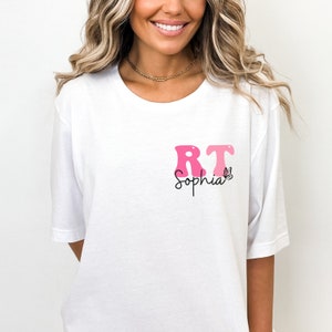 May include: White t-shirt with a pink and white "RT" graphic and the name "Sophia" written in cursive below. A small butterfly graphic is next to the name.