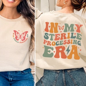 Puede incluir: Una sudadera beige con el texto "In My Sterile Processing Era" en una fuente retro colorida en la parte posterior. La parte delantera de la sudadera tiene una mariposa rosa con el mismo texto en un círculo.