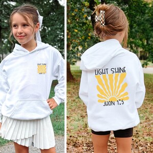 May include: White hoodie with a front pocket and hood. The front features the text "LIGHT SHINE" above a yellow sunburst design and the text "JHON 1:5". The back of the hoodie has the same design. The hoodie is worn by a person.