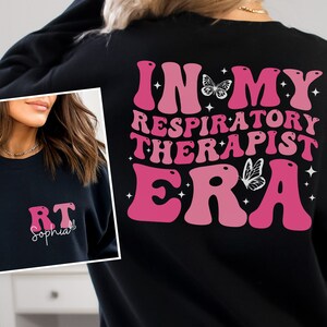 May include: Black sweatshirt with a pink and white graphic that says "In My Respiratory Therapist Era" with butterflies. The front of the sweatshirt has the letters "RT" and the name "Sophia" in white.
