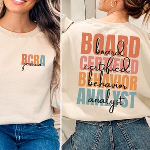 Op de afbeelding: Beige sweatshirt met de tekst "BCBA jessica" in kleurrijke letters op de voorkant. De achterkant van de sweatshirt toont de woorden "BOARD CERTIFIED BEHAVIOR ANALYST" in een gestapeld ontwerp, met behulp van oranje, roze, blauwe en zwarte letters.