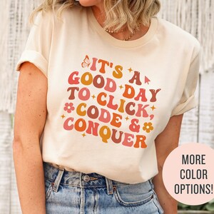 Könnte beinhalten: Cremefarbenes T-Shirt mit dem Text "IT'S A GOOD DAY TO CLICK, CODE & CONQUER" in Retro-Schrift in Orange, Rosa und Pfirsich. Ein Schmetterling und eine Blume sind im Design enthalten. Das Shirt hat auch einen "MORE COLOR OPTIONS!" Button.