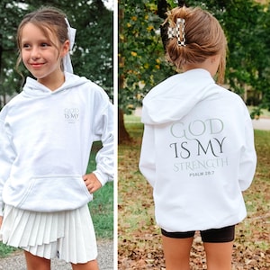 May include: A white hoodie featuring the text "GOD IS MY STRENGTH" in green and grey, with "PSALM 28:7" underneath. The hoodie is worn by a person, paired with a white pleated skirt and black shorts.