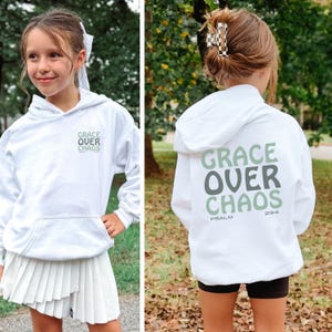May include: A white hoodie featuring the text "GRACE OVER CHAOS" in green and grey. The front and back of the hoodie display the text, with "PSALM 23:4" also printed on the back. The hoodie has a hood and a front pocket.