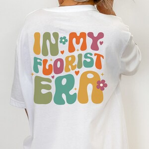 Op de afbeelding: Wit T-shirt met de tekst "In My Florist Era" in kleurrijke, retro-stijl letters. De tekst is versierd met kleine hartjes en bloemen.
