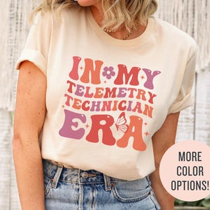 Könnte beinhalten: Ein weißes T-Shirt mit einer Grafik im Retro-Stil, auf dem "In My Telemetry Technician Era" mit einem rosa-orangenen Farbschema und einer Schmetterlingsgrafik steht.