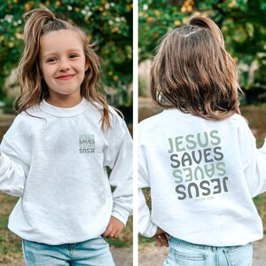 May include: A white sweatshirt featuring the text "JESUS SAVES" in shades of green and grey. The sweatshirt is worn by a young person with their hair tied up. The text is printed on both the front and back.