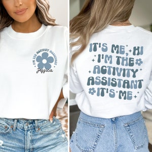 May include: White sweatshirt with a blue flower and the text "I'm the Activity Assistant Jessica" on the front and the text "It's Me, Hi I'm the Activity Assistant It's Me" on the back.