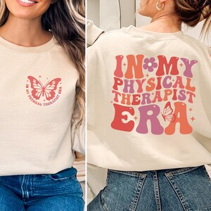 May include: A beige crewneck sweatshirt featuring a small red butterfly graphic on the front left chest with the text "IN MY PHYSICAL THERAPIST ERA". The back displays the same text in large, wavy, multi-colored red and pink letters with a butterfly.