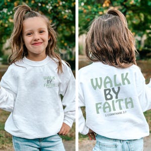 May include: A white sweatshirt featuring the text "WALK BY FAITH" in green and grey. The sweatshirt is worn by a young person with long hair. The text "2 Corinthians 5:7" is printed on the back of the sweatshirt.